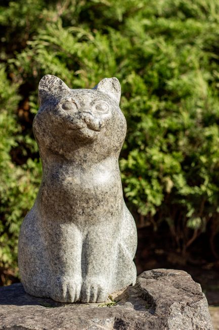 A weathered stone sculpture of a cat with a rounded face, pointed ears, and a smiling expression, seated on a flat rock surface outdoors. The sculpture has a smooth surface with some natural texture a