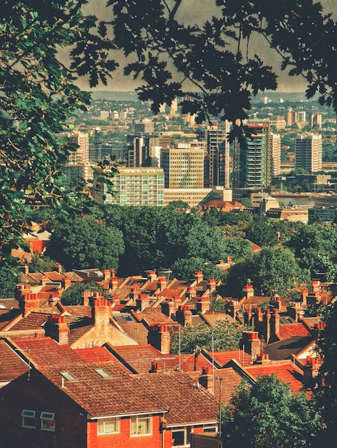 Aerial view of a residential area with rows of red brick houses featuring pitched roofs and chimneys, surrounded by abundant green trees. In the background, there is a cityscape with high-rise office